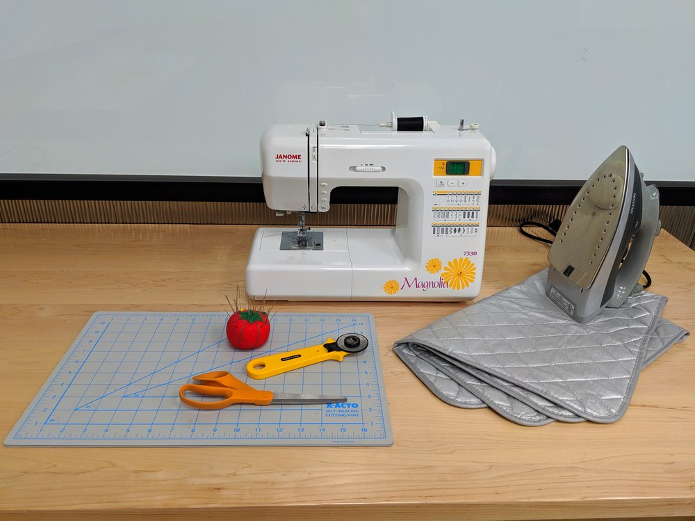 photo of sewing tools, including a sewing machine, scissors, roller cutter, cutting mat, and iron